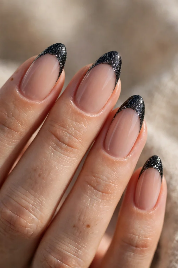 Close-up of almond-shaped nails with a nude base and sparkling black glitter French tips catching the light.