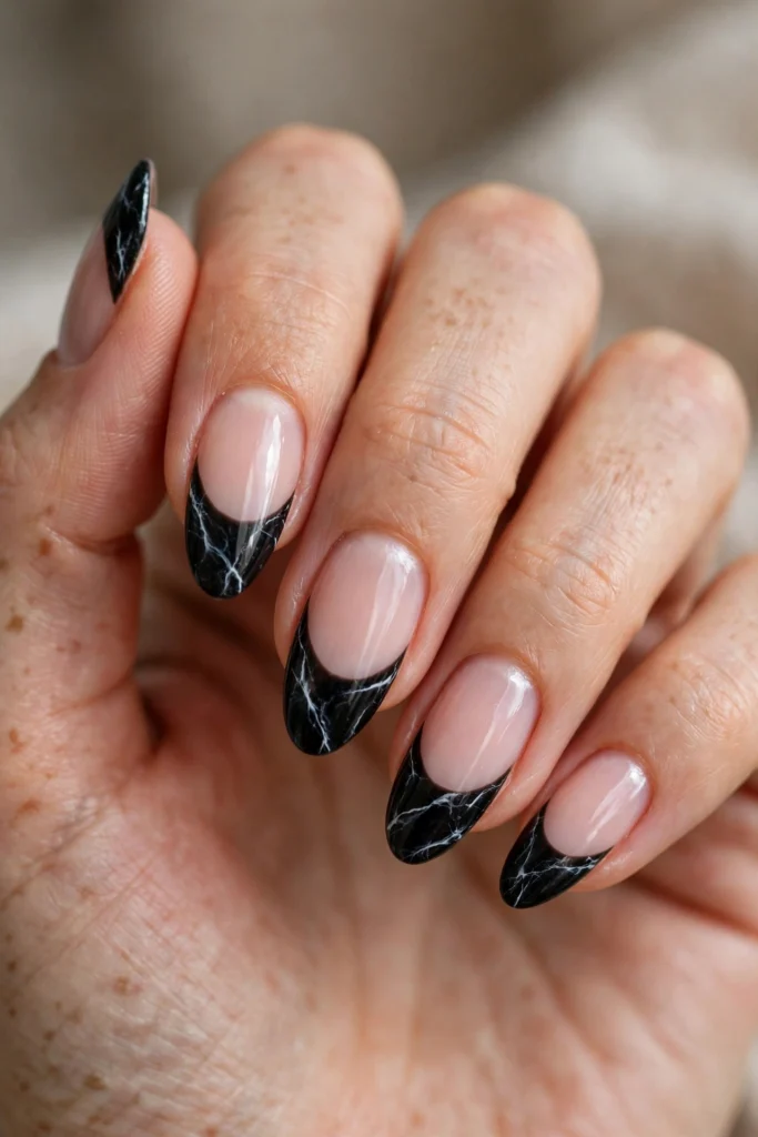 Close-up of almond-shaped nails with a nude base and black marble-effect French tips featuring fine white veining.