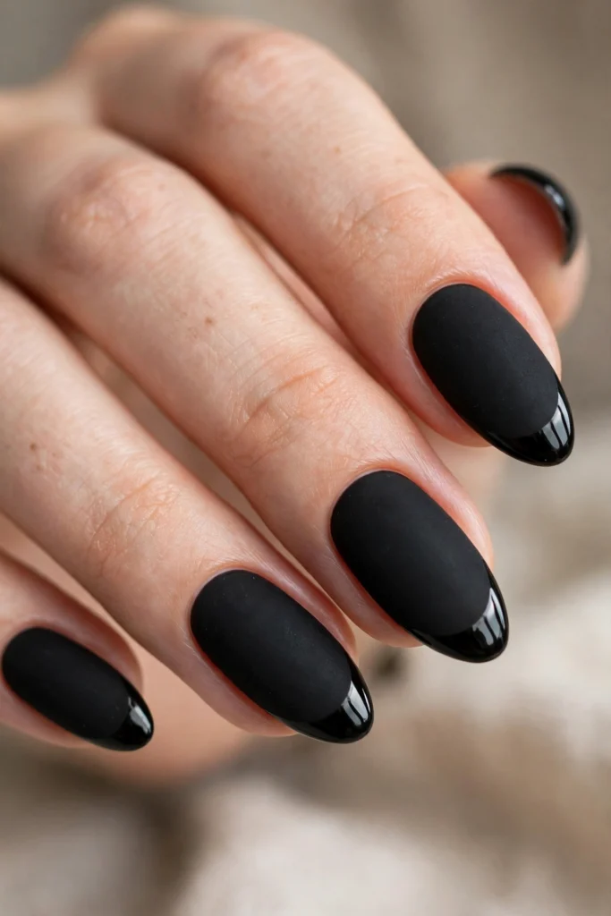 Close-up of almond nails with a flat matte black base and high-shine glossy black French tips showing clear texture contrast, portrait close-up with softly blurred background.