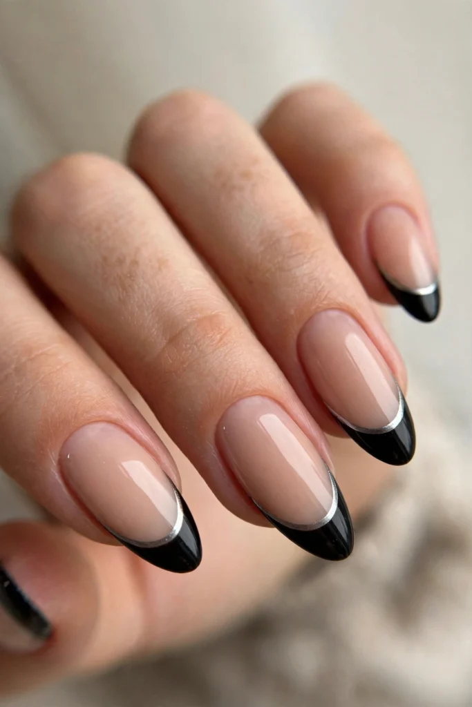 Close-up of almond-shaped nails with a sheer nude base, glossy black French tips, and a thin silver chrome line separating the black tips from the nude base.