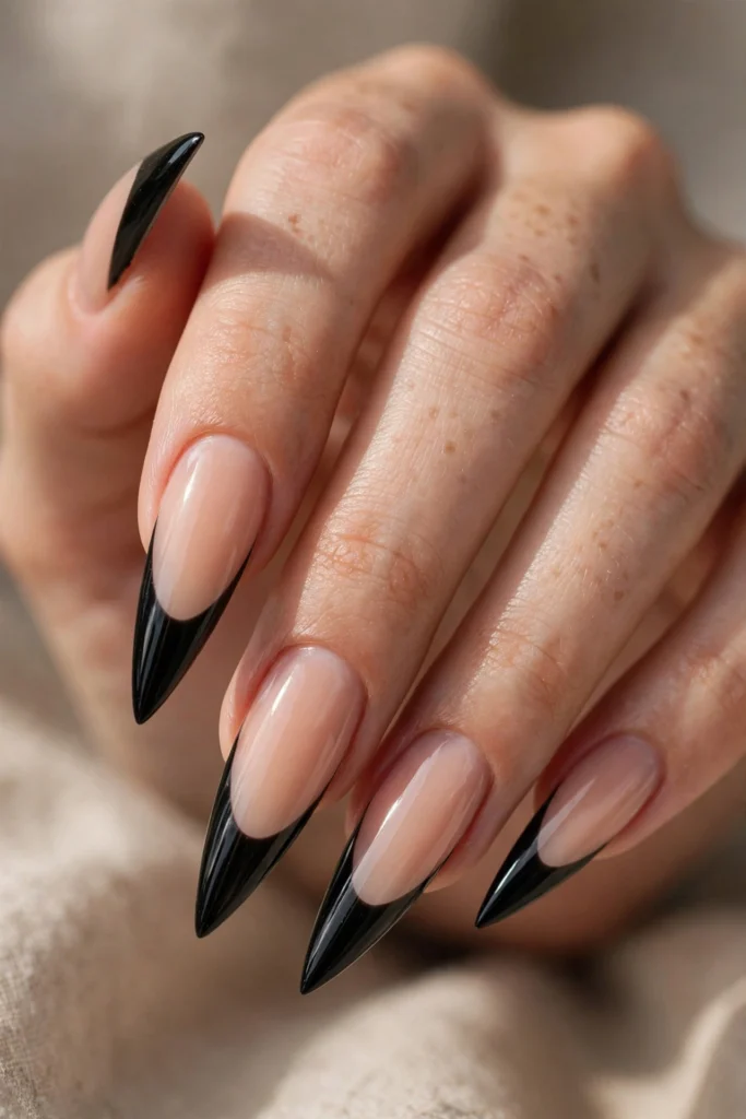 Close-up of a hand with long stiletto nails featuring glossy black French tips over a nude base, with a dramatic pointed silhouette.