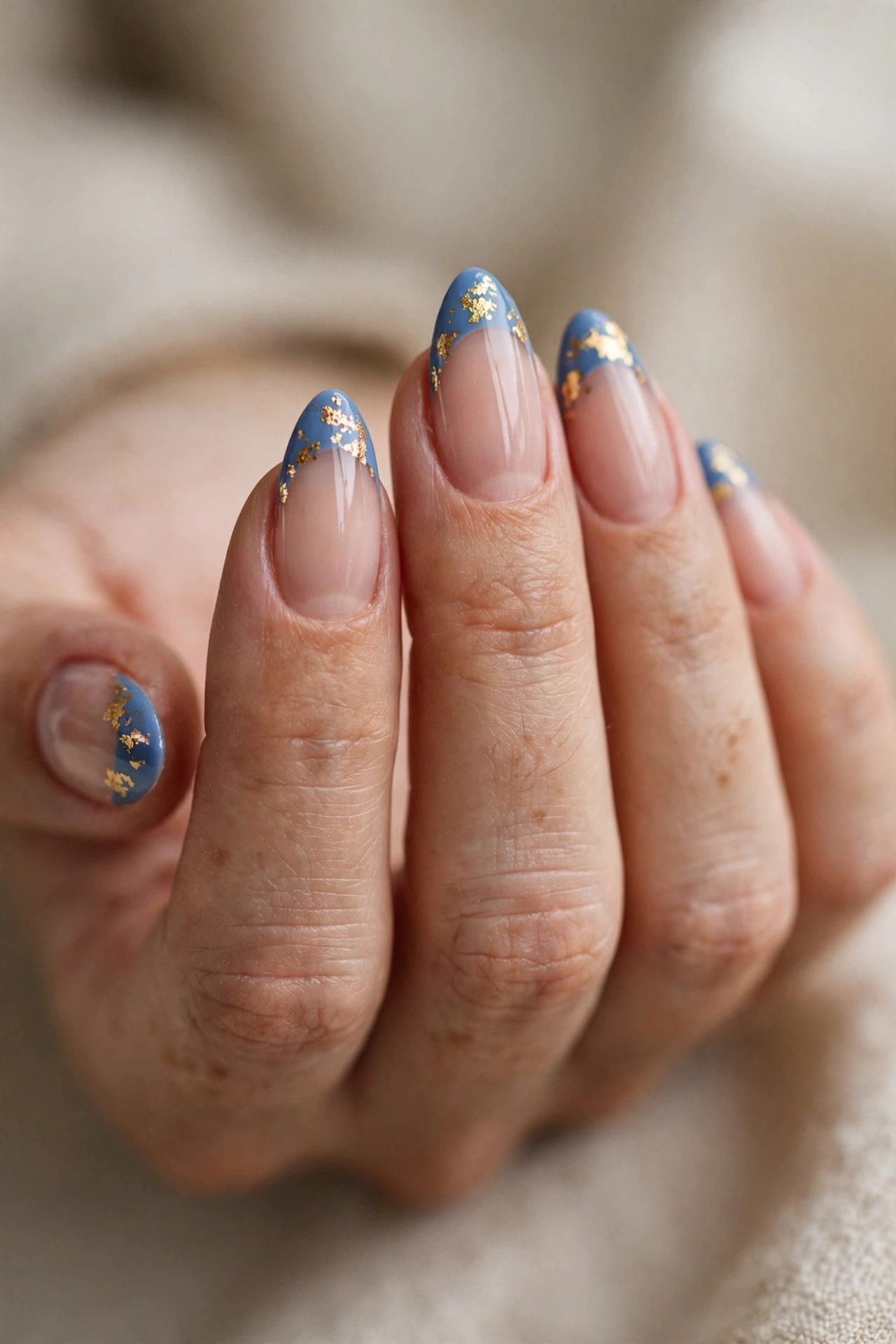 Close-up of almond nails with slate-blue French tips flecked with gold foil flakes.
