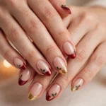 Christmas Eve French tip nails with cranberry, deep red, and gold shimmer tips and tiny holly accents on a sheer nude base, shown on a woman's hands against a warm white background with soft candlelight.