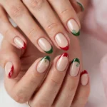 Christmas French tip nails with red and green tips and tiny snowflake art on a nude base, shown on a woman's hands against a white and soft pink background.