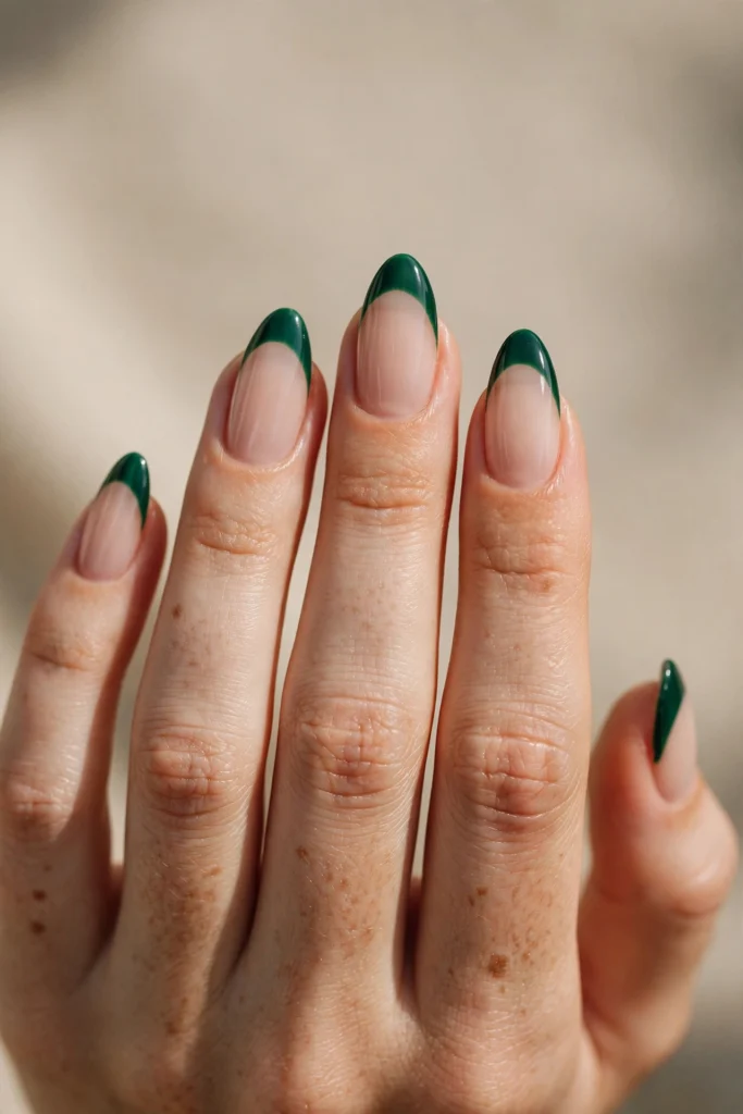 Close-up of almond nails with glossy deep emerald green French tips over a sheer nude base.
