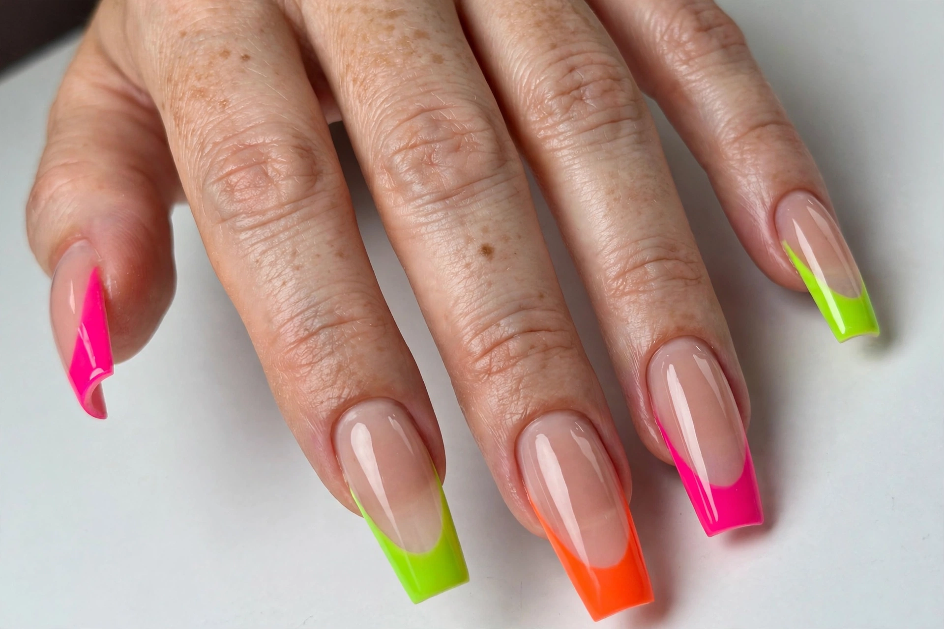 Coffin French tip nails with bright neon tips in hot pink, lime and orange on a sheer nude base, shown on long coffin nails.