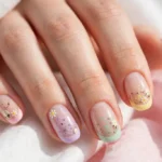 Easter French tip nails with soft pastel tips in baby pink, lilac, mint and butter yellow with tiny daisy and speckled egg art on a sheer nude base, shown on a woman's hands against a white and soft pink background.