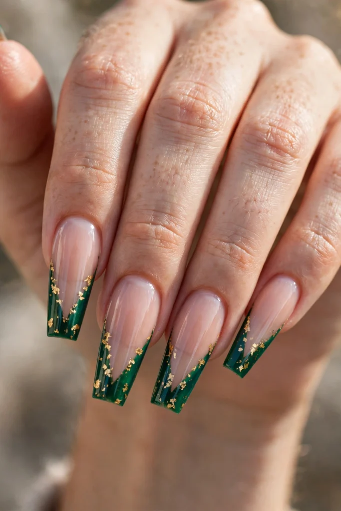 Long coffin nails with sharp V-shaped emerald French tips scattered with delicate gold foil flecks over a sheer nude base.