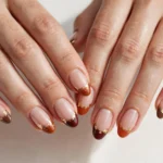 Fall French tip nails with burnt orange, burgundy, and chocolate tips and gold foil accents on a sheer nude base, shown on a woman's hands against a white and soft beige background.