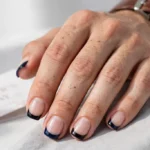 Father's Day French tip nails with deep navy and black tips and a clear glossy finish on a natural base, shown on a man's hands with short, neatly groomed nails against a white and grey background.