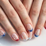 Fourth of July French tip nails with red, white and blue tips, tiny stars and glitter fireworks art on a sheer nude base, shown on a woman's hands against a white and light blue background.