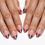 Fourth of July weekend French tip nails with red, white, and navy blue tips and tiny silver star accents on a sheer nude base, shown on a woman's hands against a white background in bright summer light.