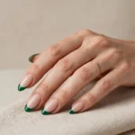 A woman's hand with almond-shaped nails wearing emerald green French tips over a soft nude base, in a wide landscape composition against a beige studio background.