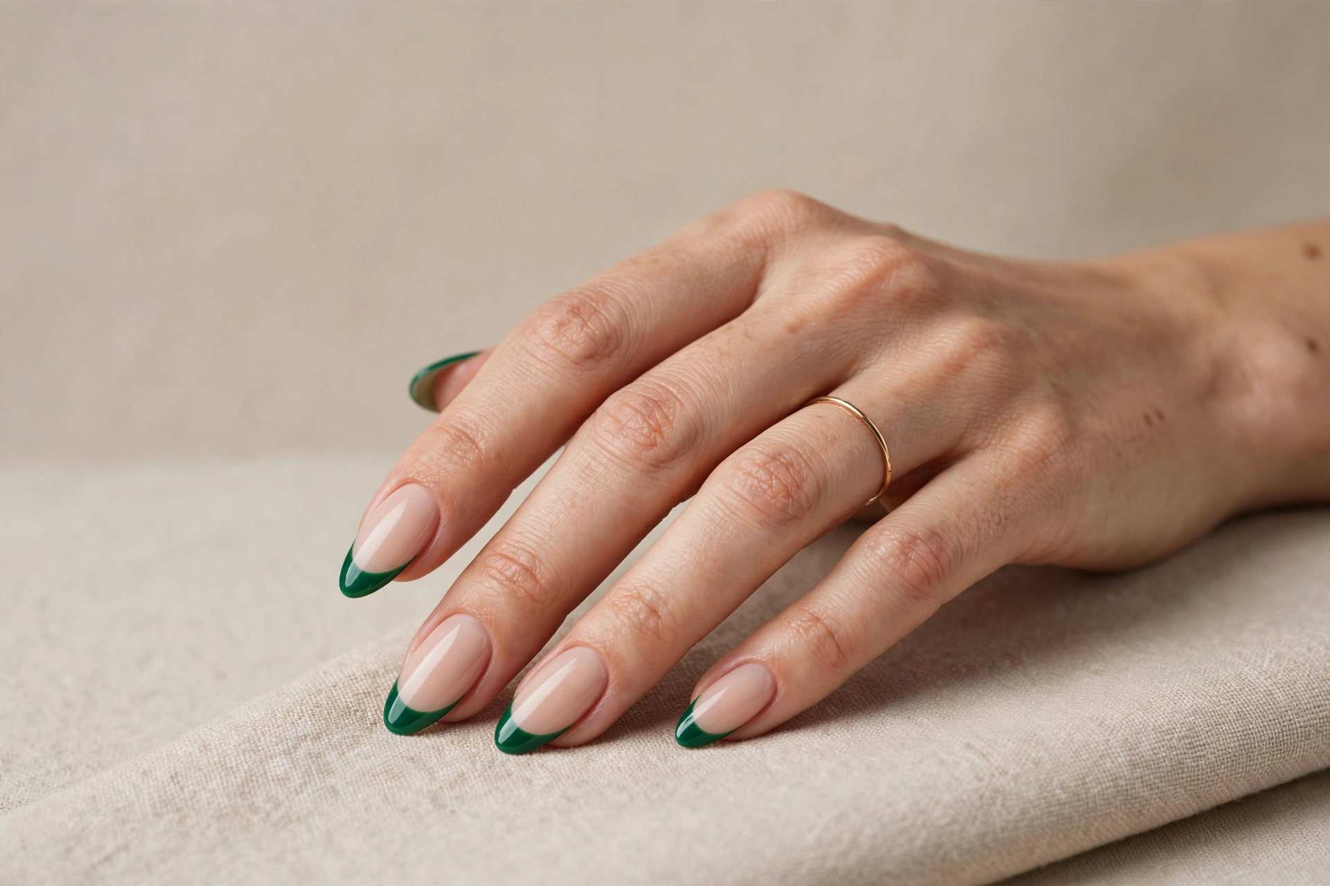 A woman's hand with almond-shaped nails wearing emerald green French tips over a soft nude base, in a wide landscape composition against a beige studio background.