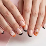 Halloween French tip nails with black and orange tips and tiny ghost and pumpkin art on a nude base, shown on a woman's hands against a white and soft pink background.
