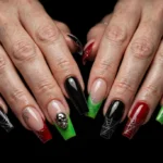Halloween party French tip nails with jet black, blood red, and slime green tips, silver spiderweb accents, and a chrome skull accent on long coffin nails, shown on a woman's hands against a black background.