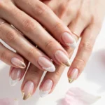 Holiday wedding French tip nails with winter white, pearl, and gold tips and subtle sparkle on a sheer nude base, shown on a woman's hands against a white background with soft winter light.