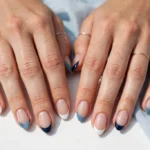 Labor Day French tip nails with crisp white and navy tips and a soft denim blue accent on a sheer nude base, shown on a woman's hands against a white and light blue background.
