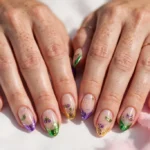 Mardi Gras French tip nails with purple, green, and gold tips, glitter accents, and tiny mask art on a sheer nude base, shown on a woman's hands against a white and soft pink background.