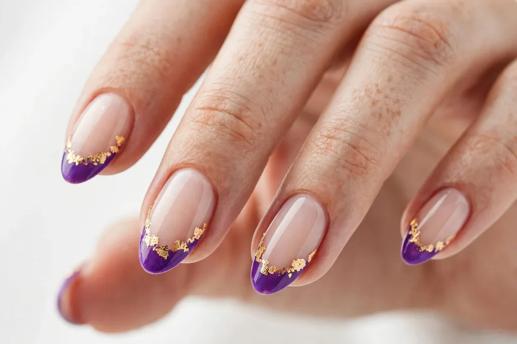 Gold Foil Mardi Gras French Tip Nails Mardi Gras French tip nails with purple tips and irregular gold foil flakes along the smile line on a sheer nude base, shown on medium almond nails.
