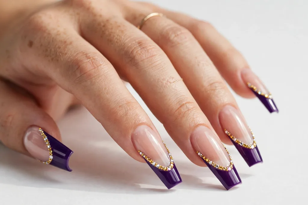 Jeweled Mardi Gras French Tip Nails Mardi Gras French tip nails with deep purple tips accented with tiny gold and crystal rhinestones along the smile line, shown on long coffin nails with a sheer nude base.