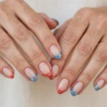 Memorial Day French tip nails with soft denim blue and coral-red tips and cream accents on a sheer nude base, shown on a woman's hands against a white and light beige background.