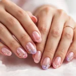 Mother's Day French tip nails with blush pink, rose and lilac tips, pearl shimmer and tiny floral art on a sheer pink base, shown on a woman's hands against a white and soft pink background.