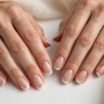 New Year's Day French tip nails with clean white tips and soft gold accents on a sheer nude base, shown on a woman's hands against a white and soft beige background.