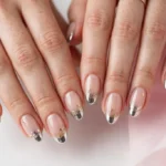 New Year's Eve French tip nails with gold glitter and silver chrome tips and tiny gold star art on a sheer nude base, shown on a woman's hands against a white and soft pink background.