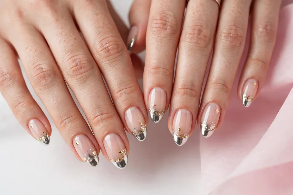 New Year's Eve French tip nails with gold glitter and silver chrome tips and tiny gold star art on a sheer nude base, shown on a woman's hands against a white and soft pink background.