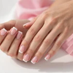 Square French tip nails with classic white tips on a sheer pink base, shown on a woman's hands against a white and soft pink background.
