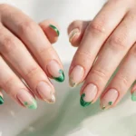 St. Patrick's Day French tip nails with emerald green, mint, and gold glitter tips and tiny shamrock accents on a sheer nude base, shown on a woman's hands against a white and soft green background.