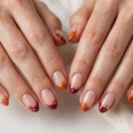 Thanksgiving French tip nails with burnt orange and cranberry tips and tiny pumpkin and fall leaf art on a sheer nude base, shown on a woman's hands against a white and soft beige background.