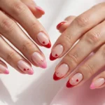 Valentine's Day French tip nails with red and pink tips and tiny heart art on a sheer nude base, shown on a woman's hands against a white and soft pink background.