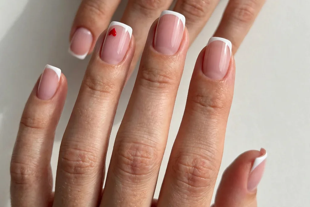 Valentine's Day French tip nails with classic white tips and one small red heart accent on the ring finger, shown on medium squoval nails with a sheer pink base.