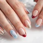 Winter holidays French tip nails with icy blue, classic red, and champagne gold tips and fine silver sparkle accents on a sheer nude base, shown on a woman's hands against a white and soft silver background.