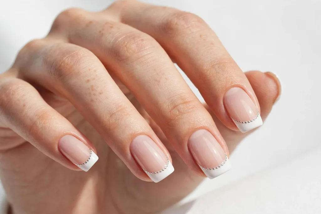 Winter holidays French tip nails with white tips edged with a row of tiny silver dots along the smile line on a sheer nude base, shown on medium squoval nails.